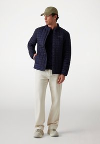 Jachetă quilted de marină cu închidere cu fermoar și buzunare laterale, purtată cu o cămașă neagră, pantaloni crem și adidași gri. Șapcă oliv.