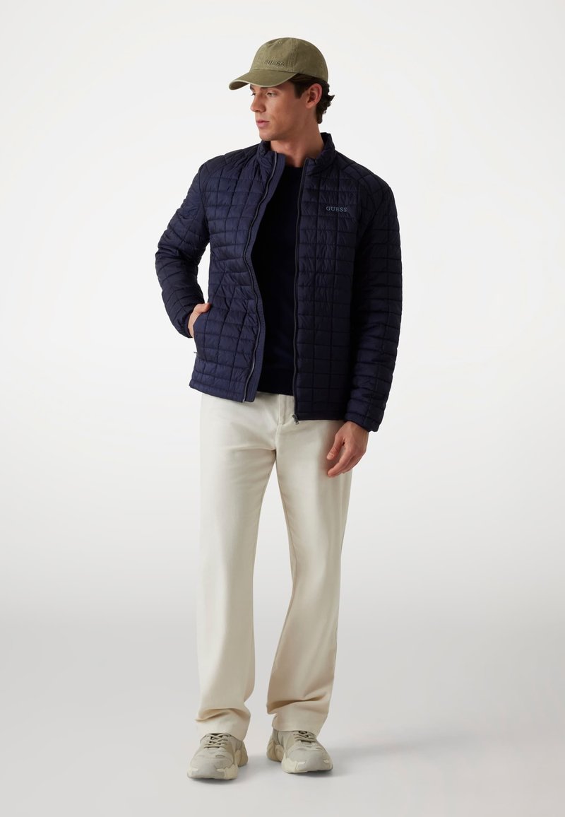 Jachetă quilted de marină cu închidere cu fermoar și buzunare laterale, purtată cu o cămașă neagră, pantaloni crem și adidași gri. Șapcă oliv.
