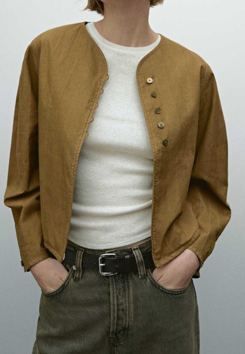 Giacca button-up marrone in tessuto strutturato, con collo rotondo, maniche lunghe e dettagli di cucitura in vista. Indossata sopra una camicia bianca.