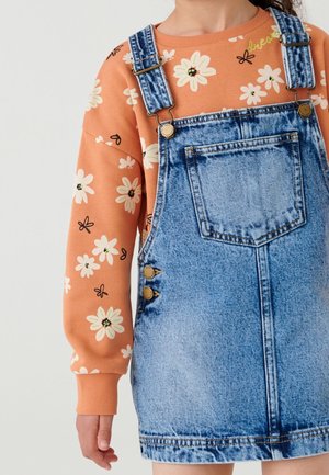 Enfant portant une chemise orange à fleurs manches longues sous une robe salopette en jean bleu avec poche avant et bretelles réglables.