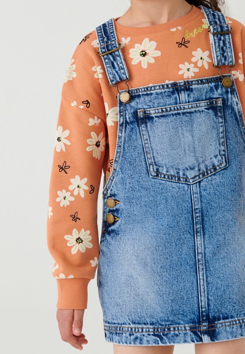 Enfant portant une chemise orange à fleurs manches longues sous une robe salopette en jean bleu avec poche avant et bretelles réglables.