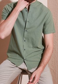 Homme assis sur un tabouret portant une chemise verte à manches courtes avec boutons et un pantalon beige, une main touchant son cou.
