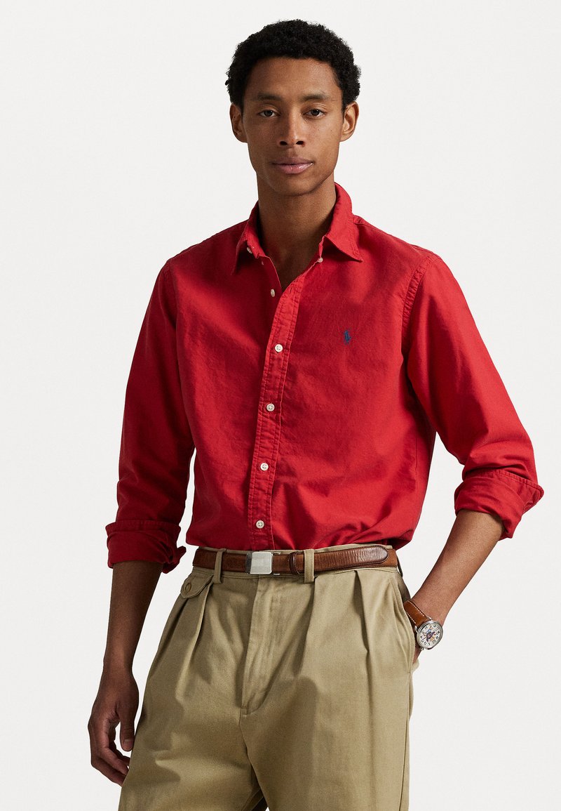 Punane puuvillane nööpidega särk väikese sinise embleemiga, üleskeeratud varrukatega, koos beežide kortsunud pükste ja pruunist vööst.