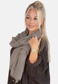 Écharpe grise clair et texturée drapée autour du cou. Présente des bords effilochés. Portée sur un haut en tissu foncé, associée à une montre en or.