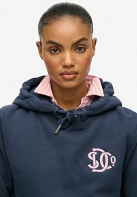 Sudadera azul marino con textura cepillada, con capucha con cordón y un logo rosa. Llevada sobre una camisa de rayas rosa claro.