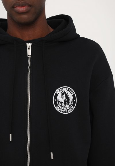 Black zip-up hoodie with a kangaroo pocket, silver zipper, and a circular white graphic logo on the left chest area. Soft fabric texture.