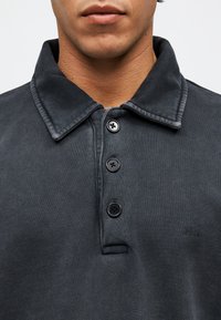 Κοντινό πλάνο ενός ατόμου που φοράει σκούρο γκρι πουκάμισο πόλο με κουμπιά και γιακά που διπλώνει, εμφανίζοντας τα χείλη και μέρος του λαιμού.