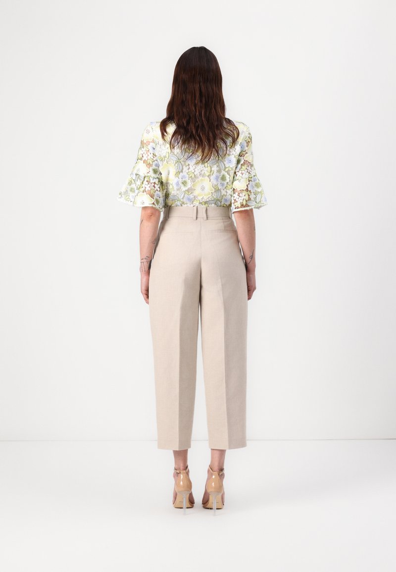 Bluse mit floralem Muster und kurzen Ärmeln in hellen Farben, kombiniert mit beige hochgeschnittenen Hosen, die einen geraden Schnitt und Knöchellänge haben.