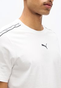 Κοντινό πλάνο ενός ατόμου που φοράει άσπρο T-shirt Puma με λογότυπο στο στήθος και χαρακτηριστικές ρίγες στους ώμους.