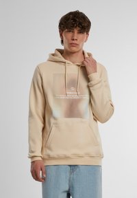 Beige hættetrøje med en frontlomme, snørret hætte og et grafikprint med hvid tekst, lavet af blødt bomuldsstof.