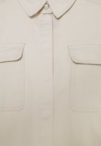 Chemise beige avec col, deux poches poitrine, et fermeture à boutons pression nacrés. La texture du tissu semble douce avec un motif de tissage visible.