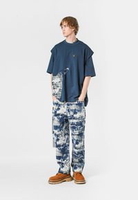 Joven con una camiseta azul marino de gran tamaño con un patrón abstracto y pantalones a juego con estampado azul y blanco, de pie con las manos en los bolsillos.