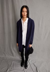 Cardigan en tricot marine avec une capuche, poches avant, associé à une chemise blanche à boutons et des leggings noirs, complété par des bottes noires à bout carré.