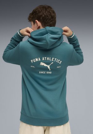 Jeune personne ajustant la capuche d'un sweat à capuche Puma Athletics turquoise avec logo et texte « Since 1948 » au dos, debout devant un fond gris uni.