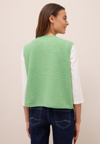 Ärmlös grön fleeceväst med en texturerad yta, rund halsringning och rak skärning, bärs över en vit långärmad tröja och mörka jeans.