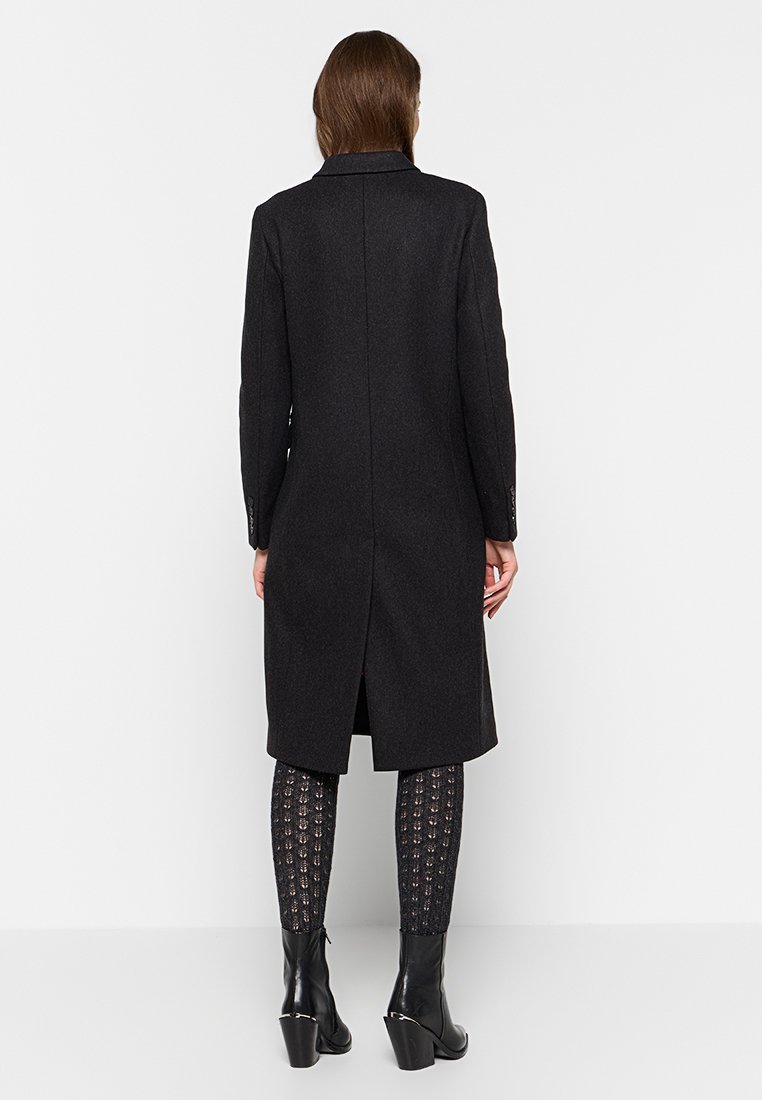 Manteau long noir en laine, avec une coupe droite, un col et une fente dans le dos. Associé à des leggings à motifs et des bottes noires à talons.
