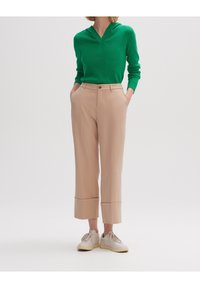 Zielona bluza z kapturem w połączeniu z beżowymi spodniami typu tailored, które mają prosty krój i złożone mankiety. Całość dopełniają białe trampki.