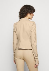 Blazer beige ajusté avec une texture lisse, col cranté et deux fentes au dos. Détails de boutons sur les manches. Design ajusté, longueur mi-cuisse.