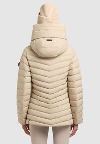 Béžová prešívaná bunda s chevronovým prešívaným vzorom, kapucňou a čiernou nášivkou na zadnej strane. Na hlave je pletená béžová čiapka.