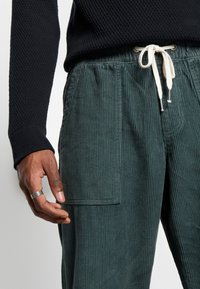 Manšestrové kalhoty ve tmavě zelené barvě s texturovaným povrchem, elastickým pasem s krémovými stahovacími šňůrkami a boční kapsou s jemným švem.