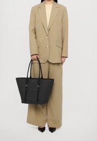 Mustat kangaskassi, jossa on teksturoitu kuvio, pitkät nahkahihnat ja hopeiset yksityiskohdat, jonka pitää päällään henkilö vaaleassa ylisuureksitussa puvussa.