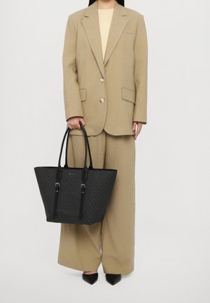 Svart tote-väska med en texturerad mönster, med långa läderhandtag och silverdetaljer, hålls av en person i en beige överdimensionerad kostym.