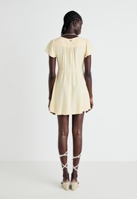Yellow short dress with flutter sleeves, fitted bodice, and flared skirt. Features a gathered back and worn with beige strappy wedge sandals.