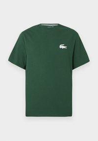 Žalias medvilninis T-shirt'as su trumpomis rankovėmis, apvaliu iškirpimu ir baltu krokodilo logotipu kairėje krūtinės pusėje. Lygi tekstūra, kasdieniškas dizainas.