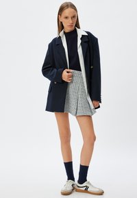 Marineblazer, hvid skjorte med krave, marineblå sweater, grå strukturerede plisserede shorts, marineblå sokker, hvide sneakers med sorte detaljer.