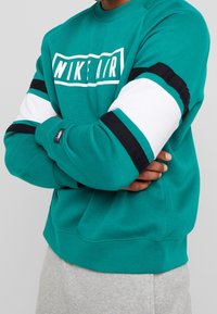 Teal sweatshirt med en vit 'NIKE AIR'-logotyp, svarta och vita randiga detaljer på ärmarna och en avslappnad passform.