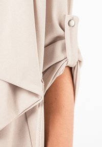 Blouse portefeuille beige clair en tissu doux et texturé. Manches retroussées maintenues par des boutons argentés et ourlet asymétrique.