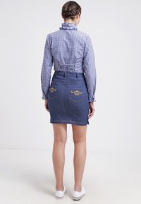 Gonna di jeans con dettagli floreali ricamati sulle tasche posteriori, dalla forma aderente e una lavaggio medio. Abbinata a una camicia a quadri blu.