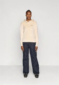 Sweat à capuche beige avec logo, associé à un pantalon cargo bleu marine avec poches latérales. Des bottes de neige noires complètent la tenue.