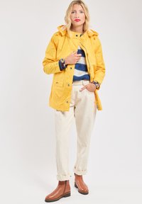 Veste imperméable jaune avec capuche, fermeture à boutons et poches avant. Portée sur un pull rayé bleu et un pantalon beige, associée à des bottes marron.