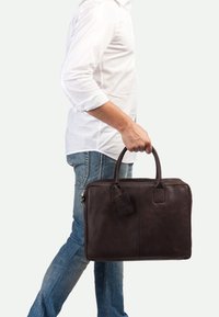Bolso para portátil de cuero marrón oscuro con forma rectangular, doble asa, una etiqueta para equipaje y una textura suave. Llevado por una persona con vaqueros.