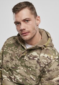 Camouflagejakke i grønne, brune og cremefarvede nuancer. Har en lynlåset hals, stor forlomme og en blød, fleeceforet hætte.