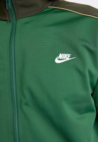 Veste zippée verte en tissu lisse, dotée d'un col montant et d'un logo Nike blanc sur la poitrine. Comprend des accents vert foncé contrastants.
