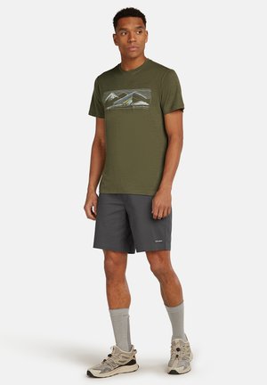 Hombre de pie vistiendo una camiseta gráfica verde oliva, pantalones cortos gris oscuro, calcetines grises y zapatos deportivos beige sobre fondo blanco.