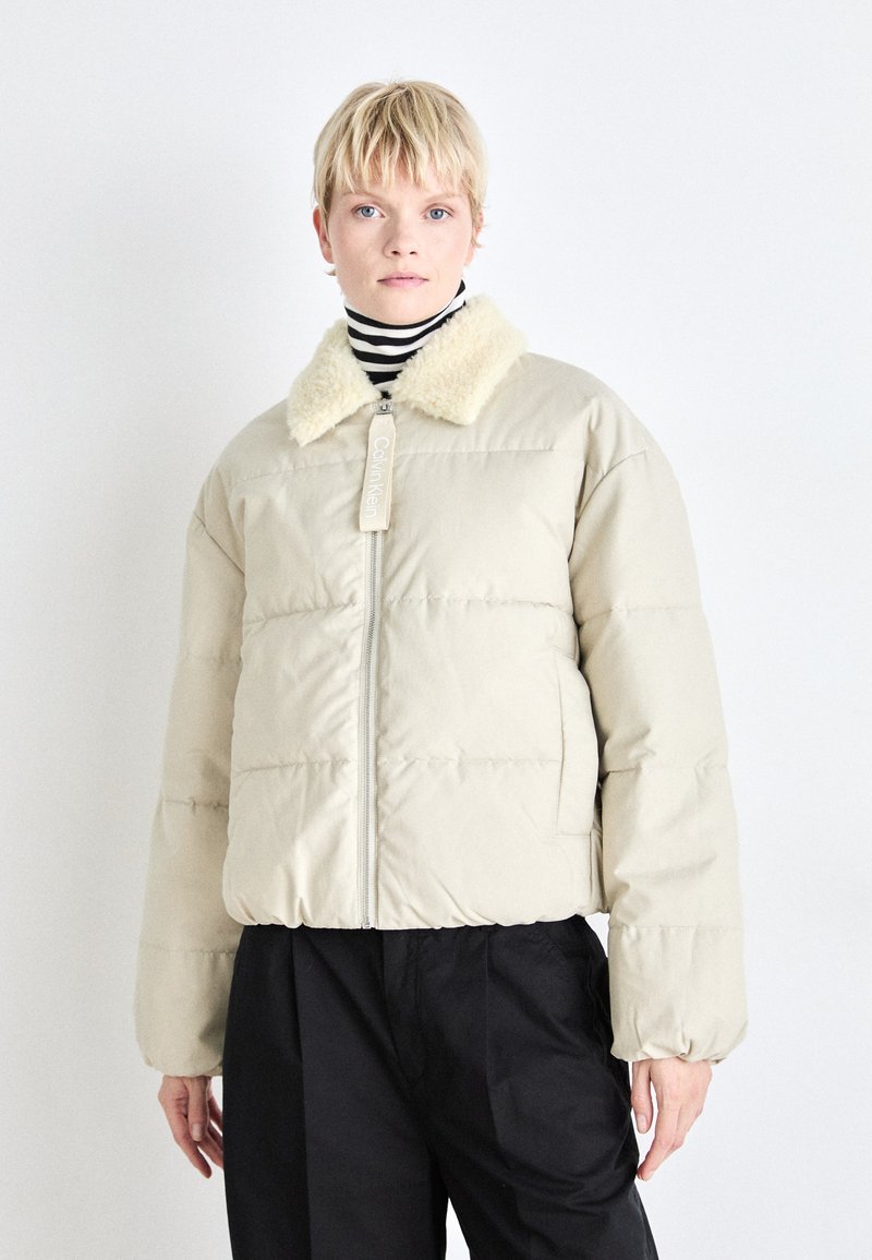 Doudoune beige avec un col en crème doux et texturé ; comporte une fermeture éclair à l'avant et une taille élastique, portée par-dessus un col roulé rayé.