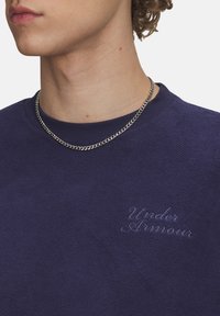 Tamsiai mėlynas tekstūruotas megztinis su apvalia kakla ir subtiliu siuvinėjimu "Under Armour" ant krūtinės. Matoma grandinėlė.
