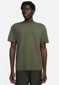 Tamsiai žalia „Nike“ marškinėliai trumpomis rankovėmis, apvaliu kaklu ir mažu užsiūtu logotipu ant krūtinės. Minkšta medvilnės medžiaga su lygia tekstūra.