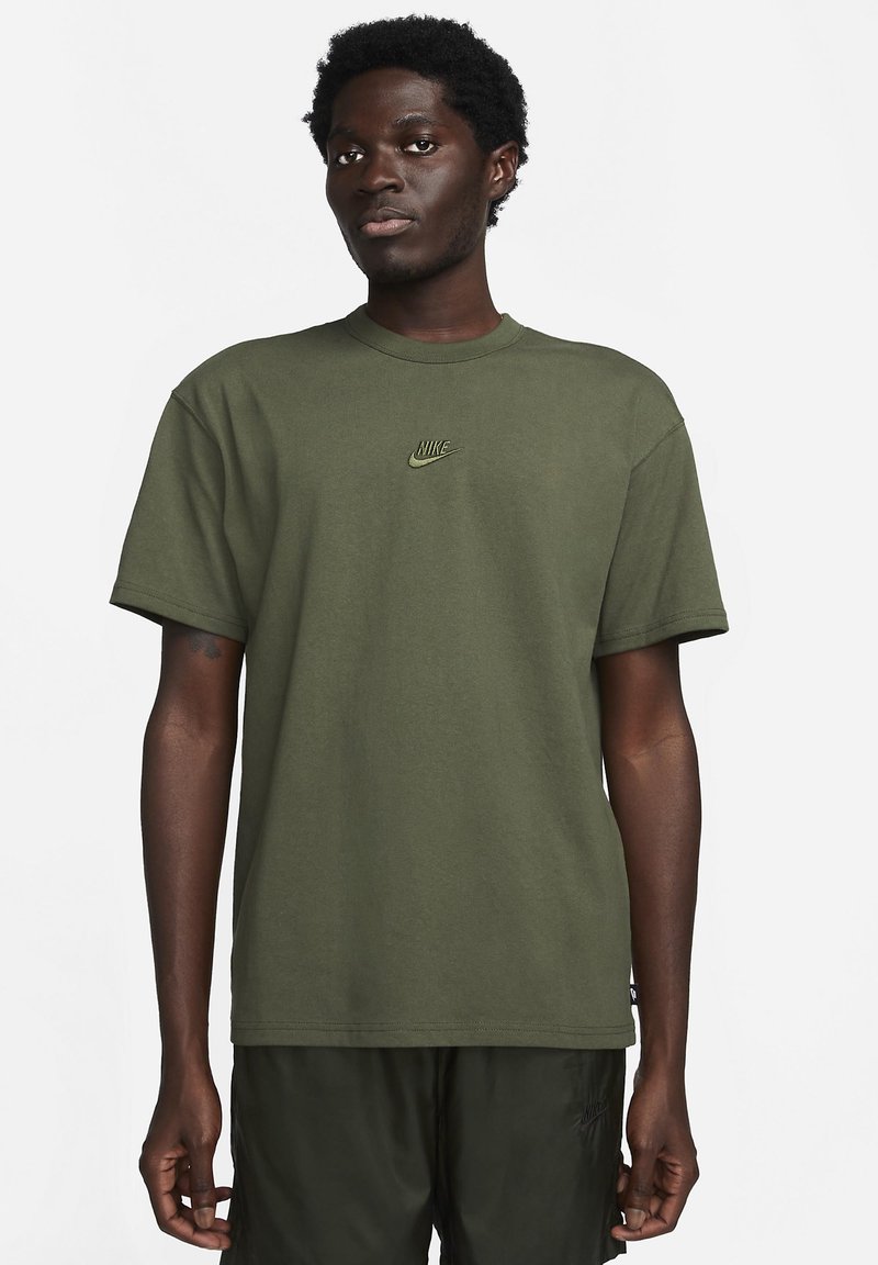Vīrietis ģērbies olīvzaļā Nike t-kreklā un tumšos šortos, stāv pretī vienkāršai baltai fona, skatoties uz priekšu.
