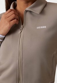 Lichtgrijze jas met ritssluiting, voorzien van een gestructureerde afwerking, een kraag en een geborduurd "GUESS"-logo in het wit op de borst.