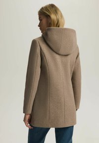 Brązowy płaszcz z kapturem, wykonany z teksturowanej tkaniny, o dopasowanym kroju i bocznych szwach, w połączeniu z niebieskimi dżinsami. Charakteryzuje się minimalistycznym designem i prostymi liniami.