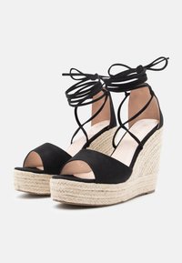 Schwarze offene Wedge-Sandalen mit geflochtenen Jute-Plattformsohlen und kreuzgekreuzten Knöchelriemen, die zu Schleifen gebunden sind.