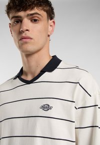 Hvid polo-shirt med marineblå horisontale striber, ribbet krave og broderet logo på venstre bryst. Glat stoftekstur.