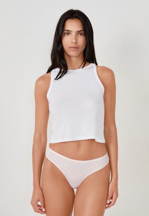 Donna con lunghi capelli scuri che indossa un top corto bianco senza maniche e mutandine rosa chiaro a vita alta, in piedi contro uno sfondo semplice.