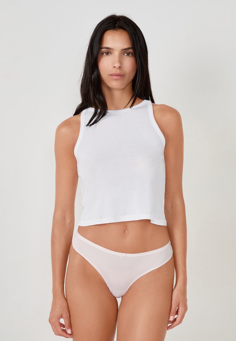 Femme aux longs cheveux foncés portant un haut court blanc sans manches et une culotte haute rose clair, debout devant un fond uni.