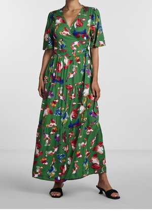 Groene wikkeljurk met veelkleurige abstracte bloemenprint, korte pofmouwen, gelaagde rok, gedragen met zwarte sandalen met hak.