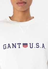 Sweatshirt blanc avec "GANT U.S.A." brodé en bleu foncé, accompagné d'une icône en rouge et bleu en dessous du texte. Tissu texturé.
